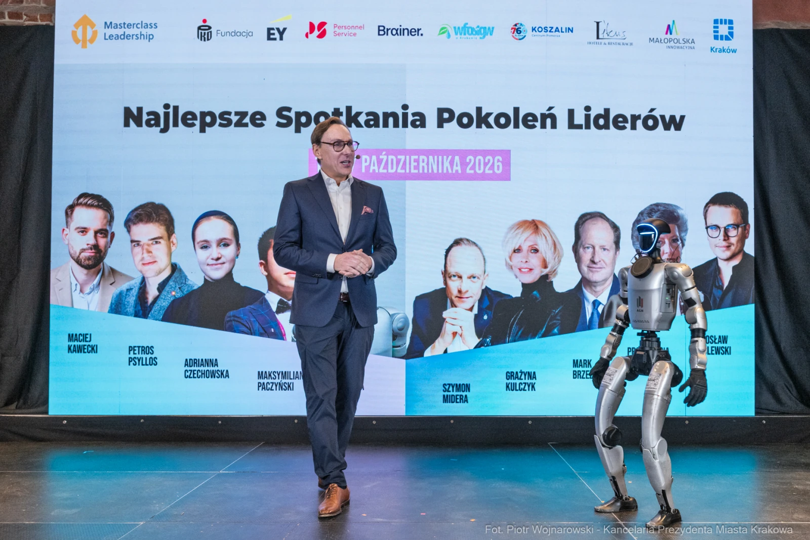Mężczyzna w garniturze prezentuje wydarzenie z robotem na scenie.  Autor: Piotr Wojnarowski