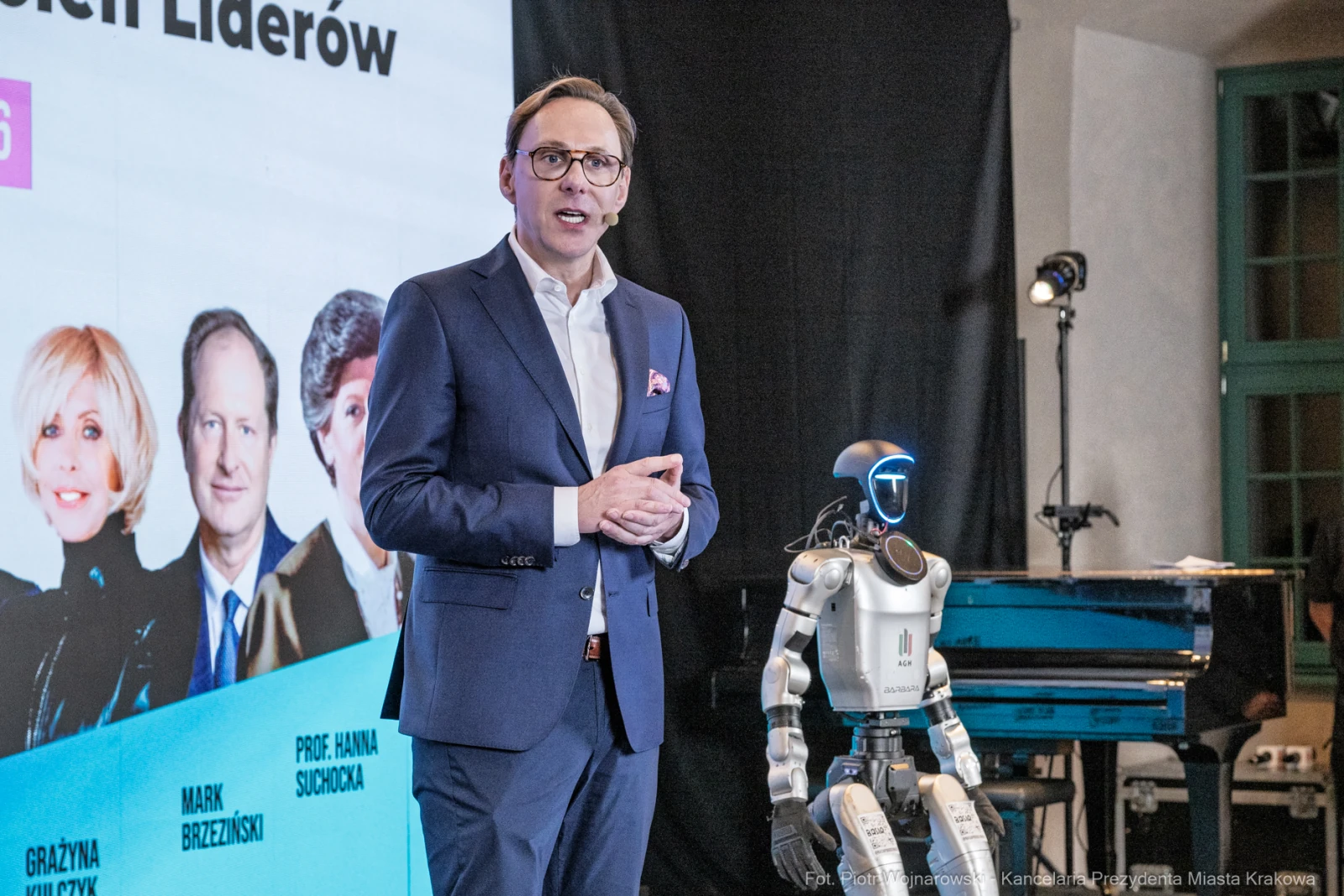 Mężczyzna w garniturze przemawia obok robota, na tle prezentacji z wizerunkami liderów.  Autor: Piotr Wojnarowski