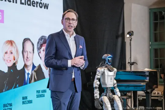 Mężczyzna w garniturze przemawia obok robota, na tle prezentacji z wizerunkami liderów.