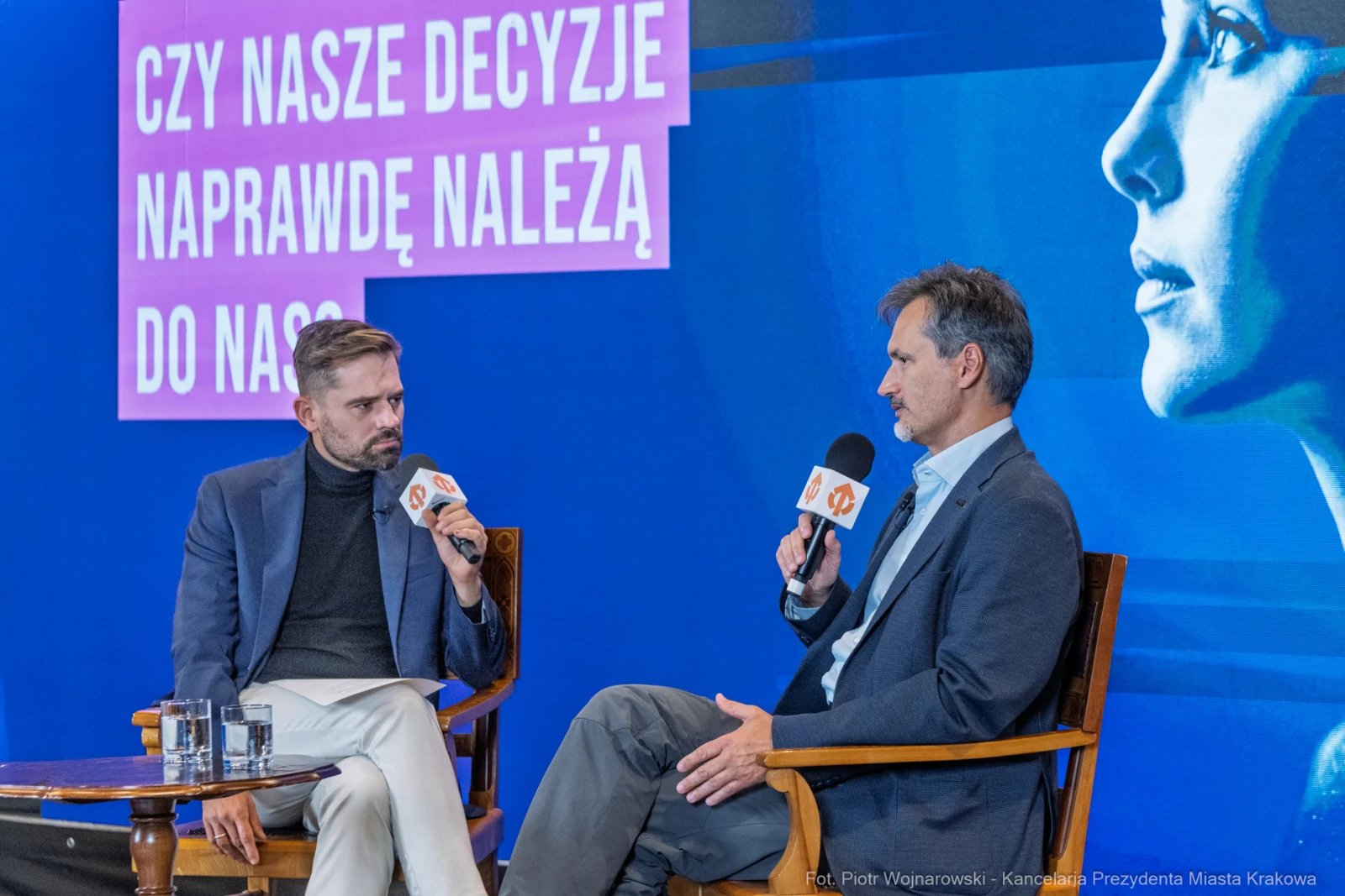 Dwóch mężczyzn rozmawia na scenie, w tle kolorowy ekran z napisem o decyzjach.  Autor: Piotr Wojnarowski