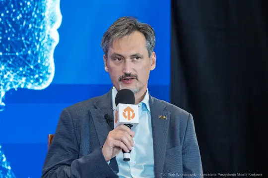 Mężczyzna z mikrofonem, prowadzący dyskusję na tle niebieskiego tła z graficznym wzorem.