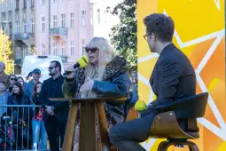 Kobieta w okularach przeciwsłonecznych rozmawia z mężczyzną przy stole, w tle tłum ludzi.