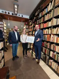 Trzy osoby w bibliotece, jedna z nich trzyma ramkę z dokumentem. W tle regały pełne książek.
