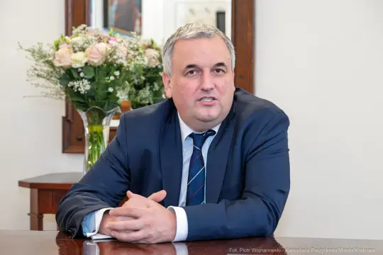 Mężczyzna w garniturze siedzi przy stole, w tle widać kwiaty w wazonie.