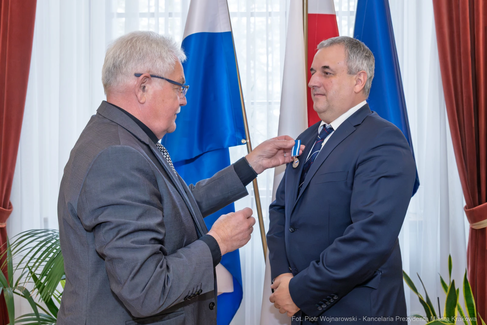 Wręczenie odznaczenia mężczyźnie w garniturze w obecności flag.  Autor: Piotr Wojnarowski