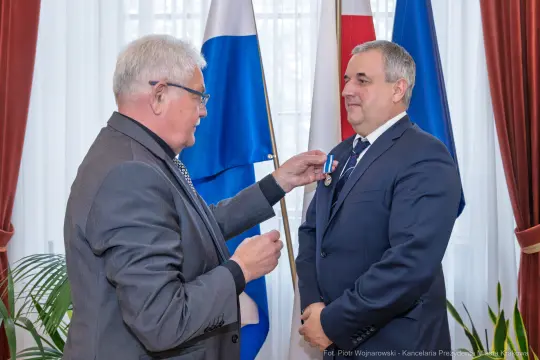 Wręczenie odznaczenia mężczyźnie w garniturze w obecności flag.