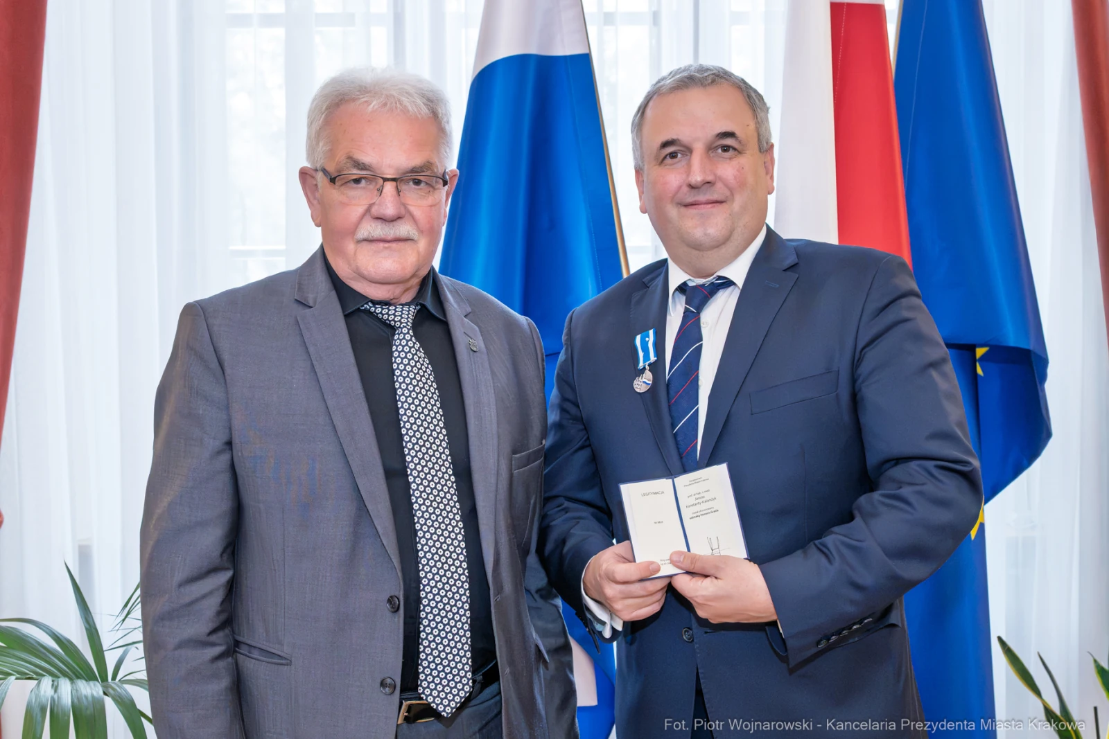 Dwaj mężczyźni w garniturach, jeden z odznaczeniem, stoją obok flag.  Autor: Piotr Wojnarowski