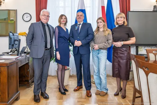 Grupa pięciu osób w biurze, w tym mężczyzna w garniturze i kobiety w eleganckich strojach.