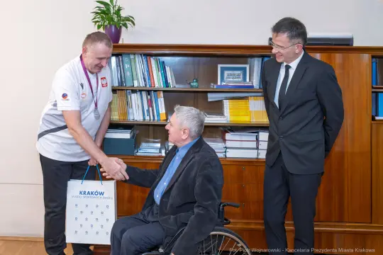 Spotkanie dwóch mężczyzn, jeden w wózku inwalidzkim, wręczenie upominku.