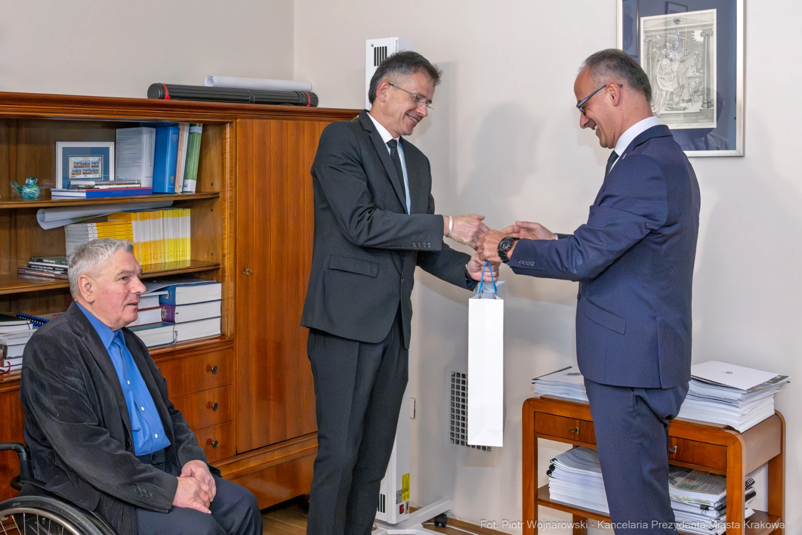 Mężczyźni w garniturach wymieniają się dokumentami, obok siedzi starszy mężczyzna na wózku inwalidzkim.  Autor: Piotr Wojnarowski