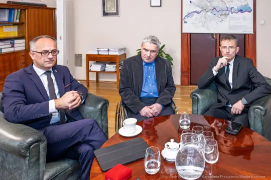 Trzech mężczyzn w garniturach siedzi w biurze, na stole filiżanki i dokumenty.