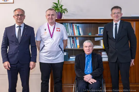 Czterech mężczyzn, w tym jeden na wózku, z medalem, stojących obok regału z książkami.