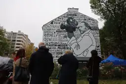 Mural z postacią trzymającą mikrofon, otoczony tekstem i datami. Grupa ludzi obserwuje dzieło.