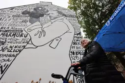Starsza kobieta z laską stoi przed muralem z portretem i tekstem.