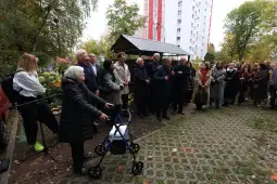 Grupa ludzi w formalnych strojach na zewnątrz, w otoczeniu zieleni i budynków.