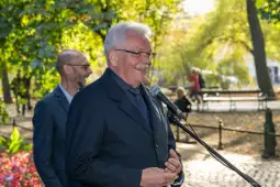 Mężczyzna przemawia do zgromadzonej publiczności w parku, w tle widać zieleń i inne osoby.