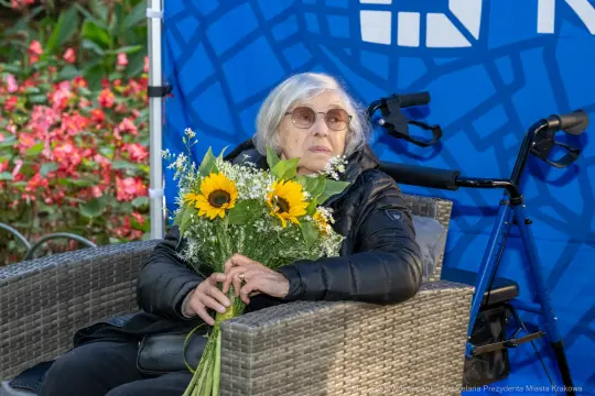 Starsza kobieta w okularach trzyma bukiet słoneczników, siedząc na krześle w parku.