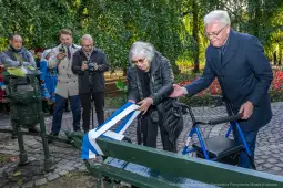 Uroczystość odsłonięcia ławki w parku, z udziałem osób starszych i obserwatorów.