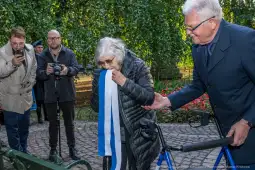 Kobieta z wstęgą w dłoni, obok mężczyzna, w tle grupa osób z aparatami.