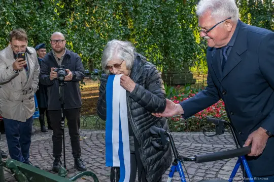 Kobieta z wstęgą w dłoni, obok mężczyzna, w tle grupa osób z aparatami.