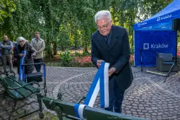 Mężczyzna przywiązuje wstążki do ławki w parku, w tle kwiaty i osoby obserwujące.