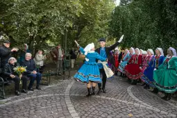 Tancerze w tradycyjnych strojach ludowych tańczą w parku, widownia obserwuje występ.