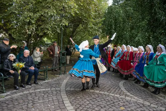 Tancerze w tradycyjnych strojach ludowych tańczą w parku, widownia obserwuje występ.