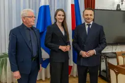 Trzy osoby w garniturach stoją obok siebie, w tle flagi.