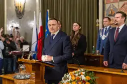 Prezentacja mężczyzny przy mównicy, w tle flagi i grupa osób w garniturach.
