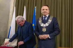 Ceremonia wręczenia odznaczeń, mężczyzna w łańcuchu prezydenckim, flagi Polski i UE w tle.