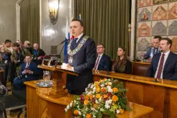 Mężczyzna w łańcuchu prezydenckim przemawia do zgromadzenia w sali obrad. W tle kwiaty i flagi.