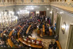 Zgromadzenie ludzi w eleganckiej sali, z mównicą i dekoracyjnym oświetleniem.