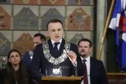 Mężczyzna w garniturze z łańcuchem na szyi przemawia do zgromadzenia. W tle widoczni inni uczestnicy.