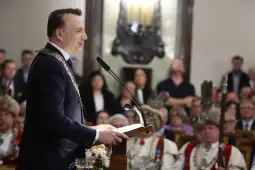 Mężczyzna w garniturze przemawia do zgromadzenia, w tle uczestnicy w tradycyjnych strojach.