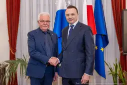 Dwaj mężczyźni uścisnęli sobie dłonie w formalnym otoczeniu, w tle flagi Polski i Unii Europejskiej.