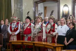 Mężczyźni w tradycyjnych strojach z odznaczeniami, stojący w sali z publicznością.
