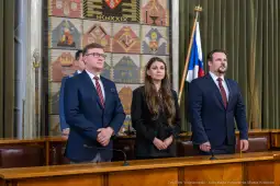 Trzech mówców stojących w sali obrad z flagami i herbami w tle.