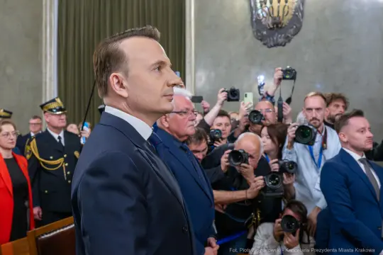 Mężczyzna w garniturze stoi na tle grupy ludzi z aparatami fotograficznymi.