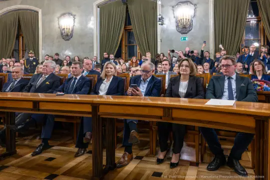 Grupa ludzi siedzących przy długim stole w sali z dekoracjami i oświetleniem.