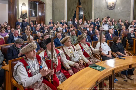 Uroczystość w sali obrad z uczestnikami w tradycyjnych strojach i publicznością na widowni.