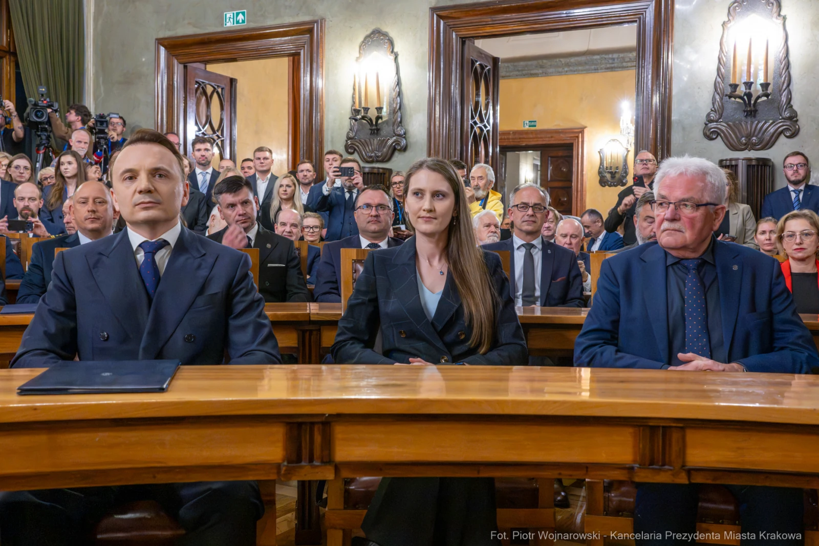 Trzy osoby siedzące przy stole w sali obrad, w tle zgromadzeni goście.  Autor: Piotr Wojnarowski