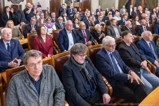 Zgromadzenie osób w sali obrad, większość w garniturach, skupionych na wydarzeniu.