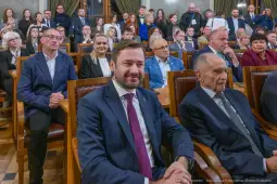 Grupa osób siedzących w sali, niektórzy uśmiechnięci, w formalnych strojach.