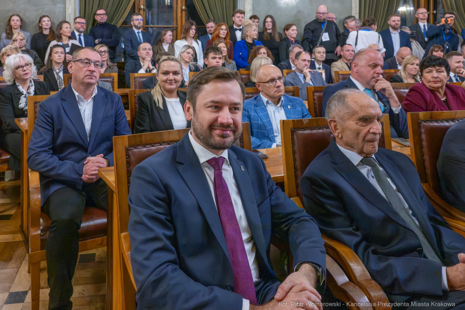 Grupa osób siedzących w sali, niektórzy uśmiechnięci, w formalnych strojach.  Autor: Piotr Wojnarowski