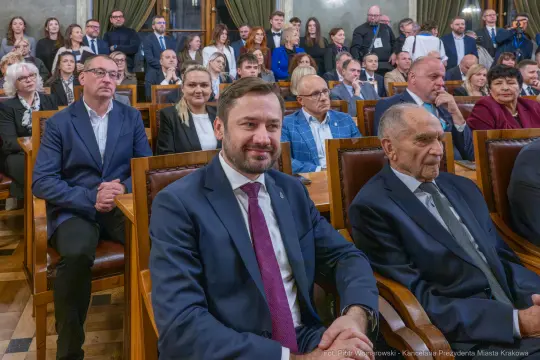 Grupa osób siedzących w sali, niektórzy uśmiechnięci, w formalnych strojach.