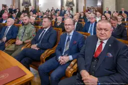 Grupa ludzi w sali, część w garniturach, część w mundurach, siedzących na krzesłach.
