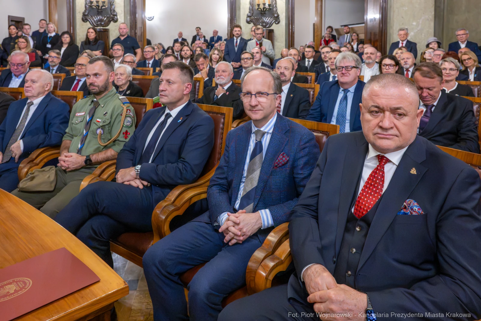 Grupa ludzi w sali, część w garniturach, część w mundurach, siedzących na krzesłach.  Autor: Piotr Wojnarowski