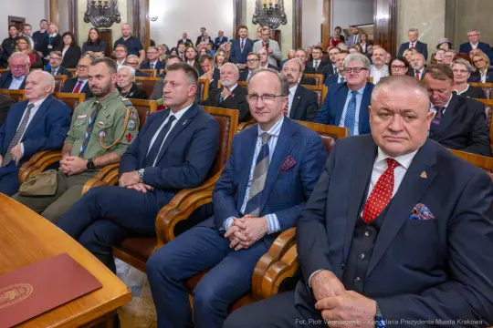Grupa ludzi w sali, część w garniturach, część w mundurach, siedzących na krzesłach.