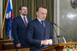 Mężczyzna przemawia z mównicy, w tle drugi mężczyzna w garniturze.