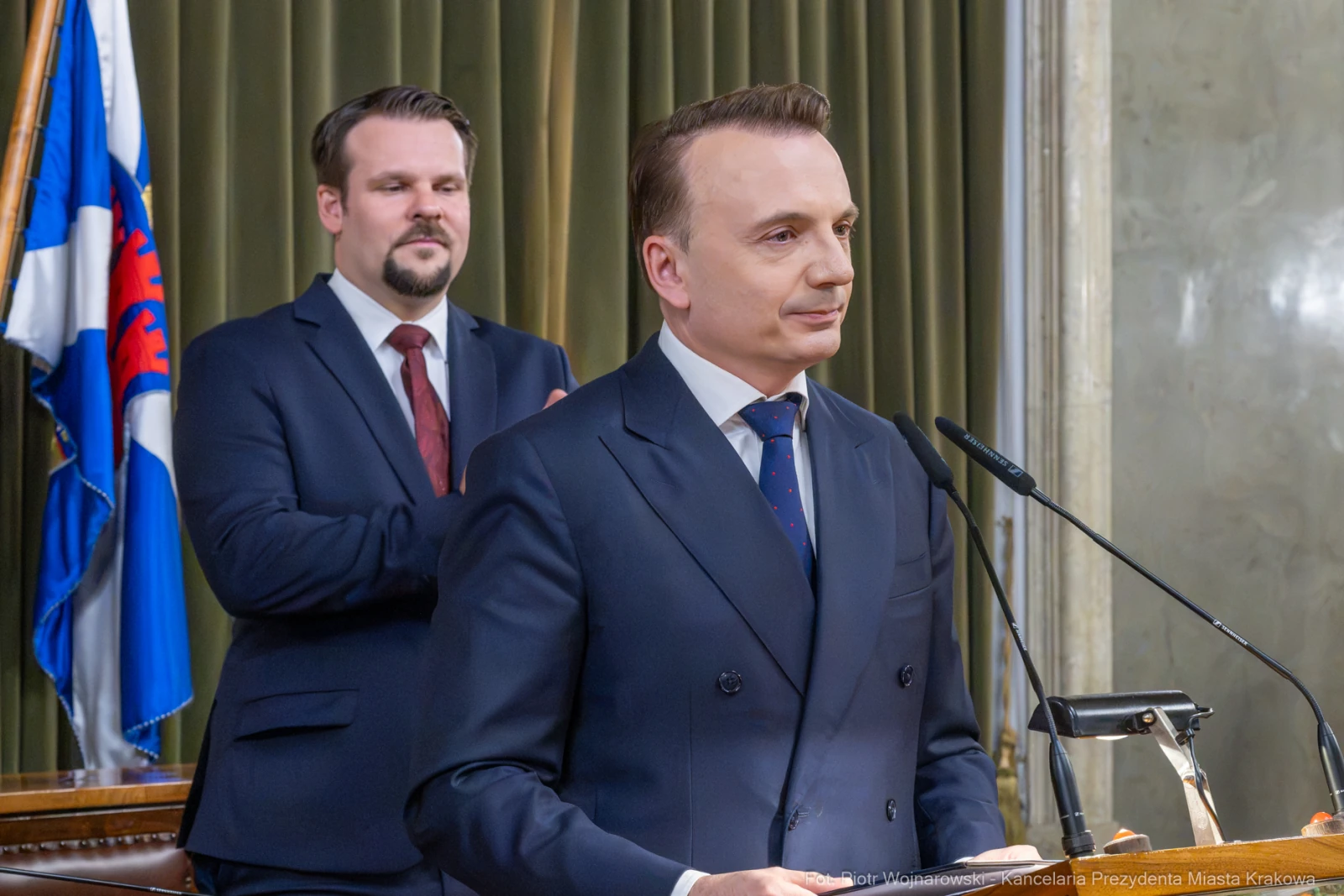 Mężczyzna w garniturze przemawia w sali obrad, drugi mężczyzna w tle aplauzuje.  Autor: Piotr Wojnarowski